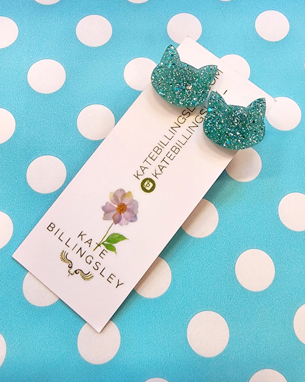 Aqua glittery cat-shaped earrings on a Kate Billingsley card with a polka dot background