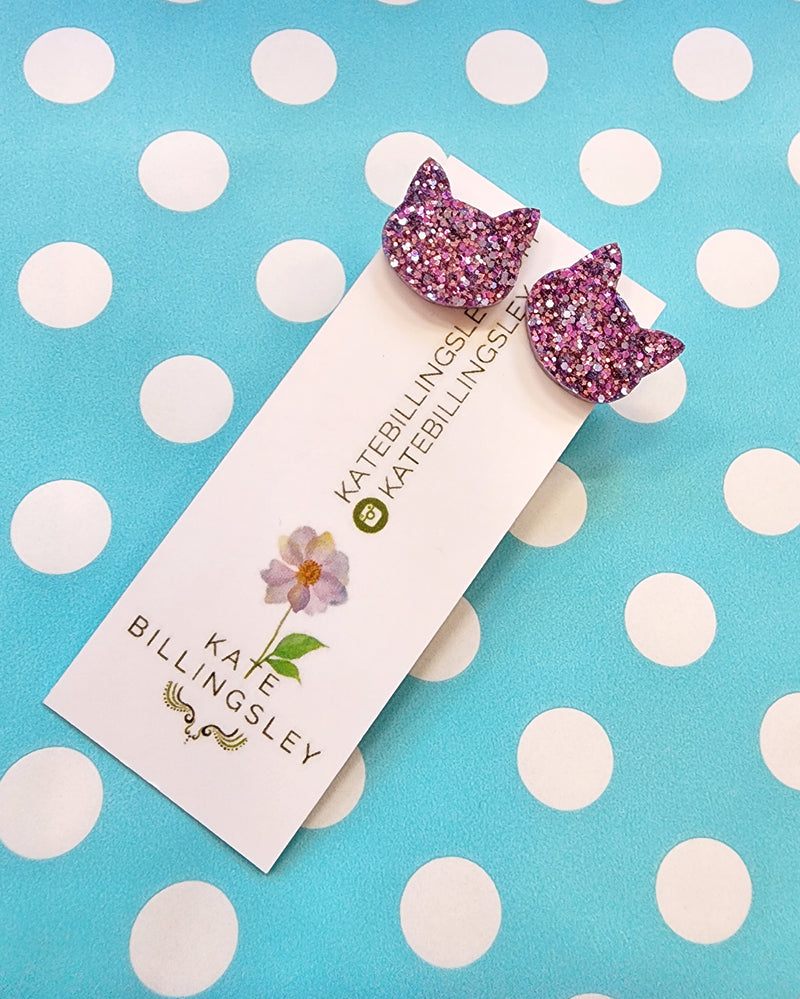 Pair of pink glittery cat-shaped earrings on a Kate Billingsley card against a polka dot background